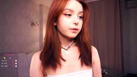 Media: Video of a young woman with fair skin, long auburn hair, and a septum piercing, wearing a white spaghetti-strap top and a silver necklace, in a dimly lit room with beige walls and a sofa.