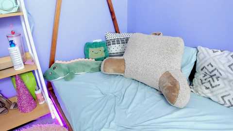 Media: Video of a cozy bedroom with a blue wall, a wooden headboard, a light blue bed, and plush toys including a green dinosaur, a beige elephant, and a white and gray pillow.