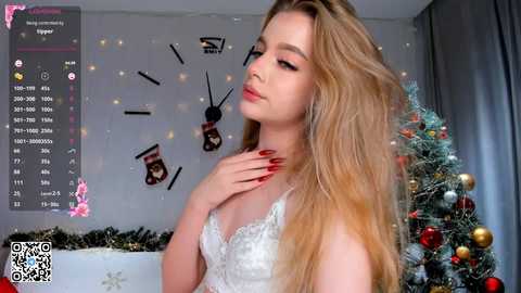 Media: Video of a young woman with long blonde hair, wearing a white lace bralette, standing in front of a decorated Christmas tree and a clock wall.