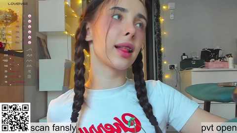 Media: Video of a young woman with fair skin, dark braided hair, wearing a white \"Walmart\" t-shirt, in a cozy room with string lights, a desk, and a green chair.