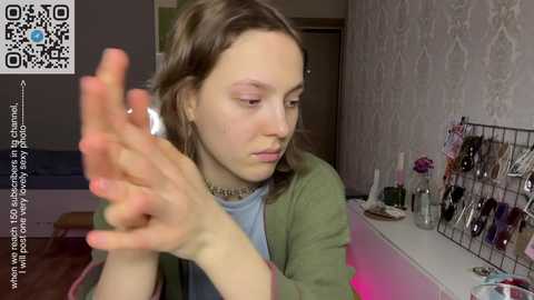 Media: Video of a young woman with light skin and shoulder-length brown hair, applying makeup in a cozy, well-lit room with floral wallpaper, makeup brushes on a table, and a QR code in the top left corner.