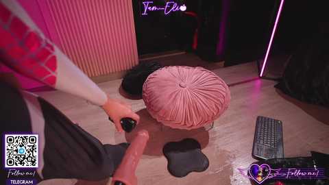 Media: Video of a virtual reality scene featuring a man's hands touching a pink, ribbed cushion on a wooden floor, with a black keyboard and a QR code visible.