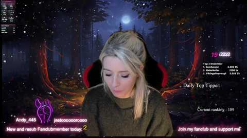 Media: Video of a young blonde woman with fair skin and light makeup, wearing a dark green sweater, sitting in a dark, forested setting with a full moon and red glow.