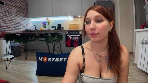 Media: Video of a fair-skinned woman with long auburn hair, wearing a gray tank top, standing in a modern kitchen with a brick wall, stainless steel appliances, and a blue \"MOSTBET\" bag on the floor.