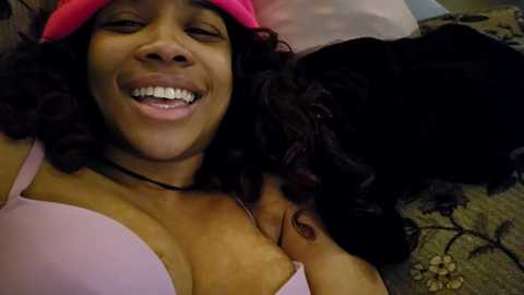 Media: Video of a joyful Black woman with curly hair, wearing a pink bra and hat, lying on a bed with a black cat beside her.