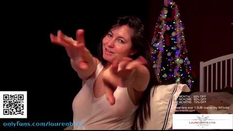 Media: Video of a smiling, long-haired woman in a white tank top, reaching out playfully. Christmas tree adorned with colorful ornaments in the background.