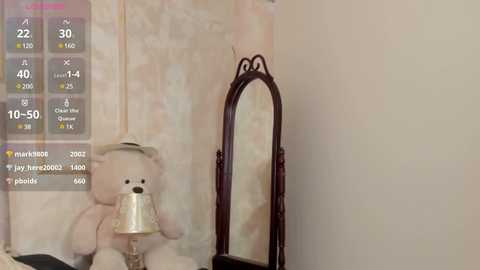 Media: A video of a cream-colored bear with a white bow, sitting on a table next to an ornate, dark wooden mirror. The background has a beige and pink floral wallpaper.