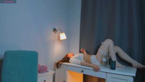 Media: Video of a young woman with fair skin, long brown hair, wearing white lingerie, lying on a white desk in a dimly lit room with blue walls and a teal chair.