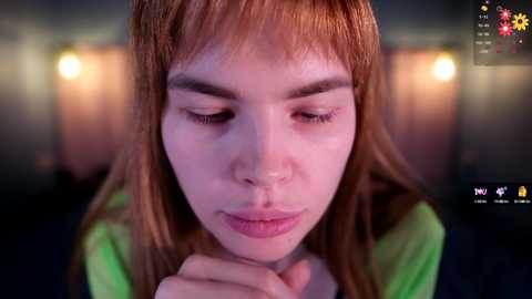 Media: Video of a young woman with fair skin, long red hair, and a green shirt, looking contemplative with her hands clasped. Background is blurred with warm, yellowish lights.