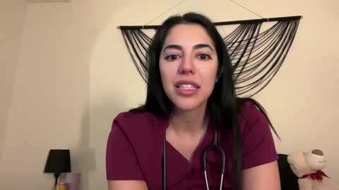 Media: A video of a Latina woman with long, black hair, wearing a burgundy scrub top, and holding a stethoscope, standing in a bedroom with beige walls and a black macram\u00e9 wall hanging.