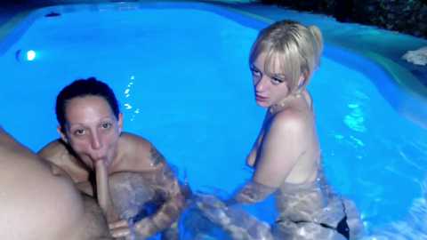 Media: A video captures a naked man and woman in a blue-tinted swimming pool. The man, with a dark complexion and muscular physique, performs oral sex on the woman, who has fair skin, blonde hair, and medium-sized breasts. The background is dark and indistinct.