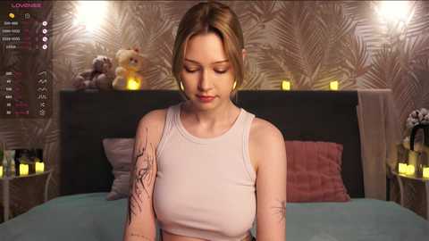 Media: Video of a young woman with light skin, brown hair, and a tattoo on her arm, wearing a white tank top, standing in a cozy, dimly-lit bedroom with a plush teddy bear, candles, and a bed with teal sheets.