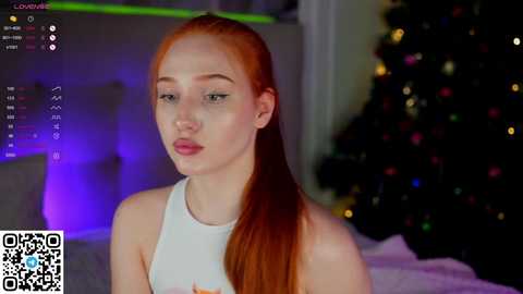 Media: Video of a fair-skinned, red-haired woman with a thin build wearing a white tank top, seated in a dimly lit room with a festive, Christmas-decorated background.