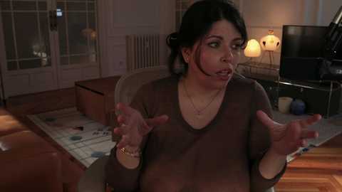 Media: Video of a middle-aged woman with dark hair in a casual brown sweater, animatedly gesturing indoors; wooden floors, vintage lamps, and a TV in the background.