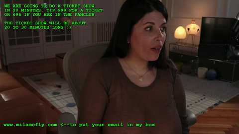 Media: A video of a woman with long black hair, wearing a brown top, seated in a dimly lit living room with a TV and lamps. Green text overlays the image with a phone conversation transcript.