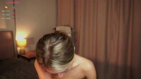 Media: Video of a nude woman with light brown hair in a bun, seated indoors with warm lighting, soft curtains, and a lit table lamp in the background.