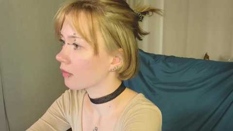 Media: A video of a fair-skinned woman with blonde hair in a bun, wearing a beige top and black choker, seated on a blue cushioned surface in a softly lit room.