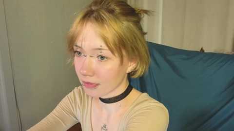 Media: Video of a fair-skinned young woman with short, light brown hair, wearing a beige top, black choker, and blue earrings, sitting on a blue cushioned chair.