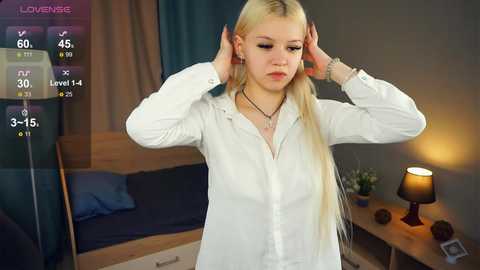 Media: Video of a young, slender, fair-skinned woman with long blonde hair, wearing a white shirt, adjusting her hair in a bedroom with a bed, nightstand, and lamp.