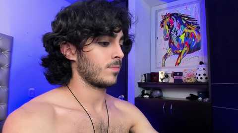 Media: Video of a shirtless man with curly dark hair and a beard, wearing a necklace, in a room with a colorful abstract horse painting on the wall, and a dark wooden dresser.