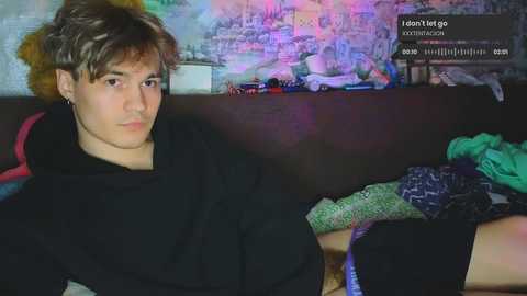 Media: Video of a young man with messy, light brown hair, wearing a black hoodie, lying on a bed with colorful pillows and a patterned blanket, in a dimly lit room with a colorful mural and gaming chat window in the background.