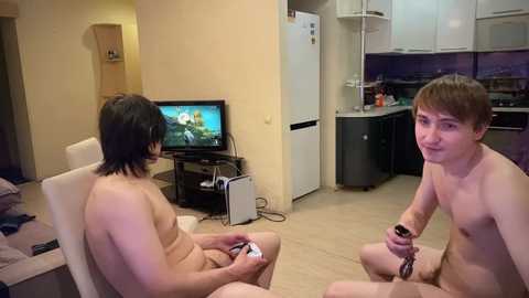 Media: Video of two young, nude, Caucasian men playing a video game on a flat-screen TV in a minimalist, beige-toned apartment kitchen.