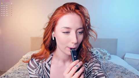 Media: A video of a fair-skinned woman with long, wavy red hair, wearing a striped shirt, holding a smartphone to her mouth, in a softly lit bedroom with white walls and a bed in the background.