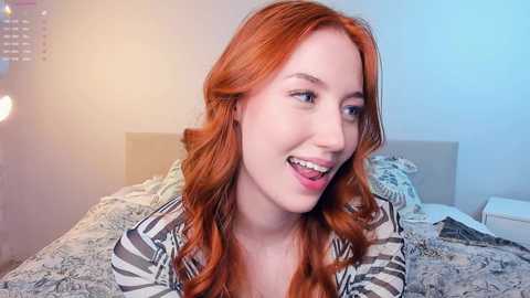 Media: Video of a young woman with long, wavy red hair, wearing a striped shirt, smiling, on a bed with patterned blankets in a softly lit, modern bedroom.