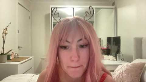 Media: A video of a fair-skinned woman with long, pastel pink hair, wearing a red bra, standing in a minimalist bedroom with white walls and a large mirror.