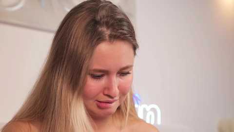 Media: A video of a fair-skinned, young woman with straight, shoulder-length blonde hair and light makeup, gazing downward with a slight smile. The background is a blurred, softly lit room with white walls and decorative elements.