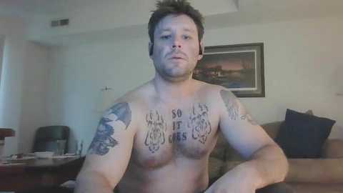 Media: Video of a shirtless, muscular, light-skinned man with short dark hair, multiple tattoos on chest and arms, sitting in a living room with beige walls, a couch, and framed art.