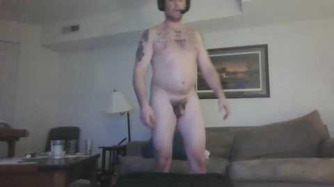 Media: Video of a naked, tattooed man with a small penis, standing in a living room with a beige sofa, a lamp, and a framed picture on the wall.