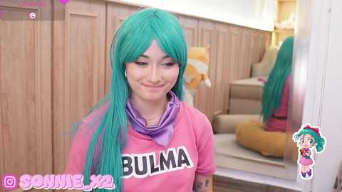 Media: Video of a smiling woman with long turquoise hair in a pink Bulma cosplay outfit, standing in a wooden-paneled room with a staircase and a large mirror.