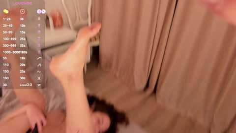 Media: A blurred video of a nude woman with medium build, lying on her back with legs raised, in a beige room with curtains.