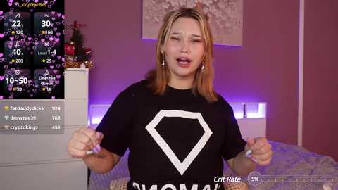 Media: Video of a young woman with light skin and blonde hair, wearing a black t-shirt with a white hexagon design. Background shows a pink bedroom with a bed, a Christmas tree, and a digital screen displaying live stats.