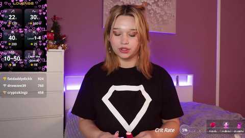 Media: Video of a young, fair-skinned woman with shoulder-length blonde hair wearing a black t-shirt with a white hexagon design, standing in a pink-painted bedroom with a bed and decorative wall art.