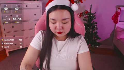 Media: Video of an East Asian woman with long black hair, wearing a red Santa hat, white T-shirt, and red lipstick, seated in a pink gaming chair, in a cozy room with a decorated Christmas tree.