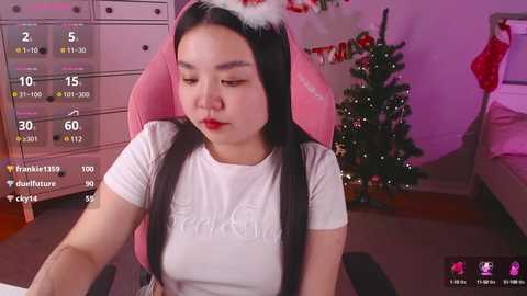 Media: Video of an Asian woman with long black hair, wearing a white Adidas t-shirt, pink Santa hat, and red lipstick, sitting in a pink gaming chair, in a festive room with a decorated Christmas tree.