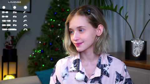 Video of a young Caucasian woman with light skin and shoulder-length blonde hair, wearing a white blouse with black dog patterns, smiling. Background features a Christmas tree, potted plants, and a fireplace.