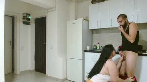 Media: Video of a man in a black tank top and no pants, standing in a modern kitchen, being orally pleasured by a woman in a white tank top.