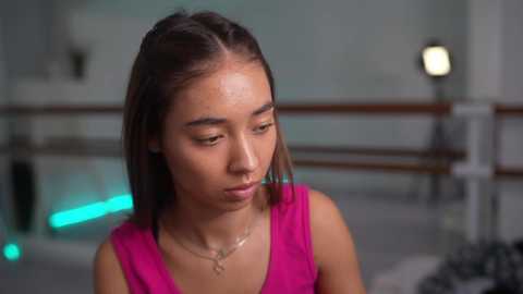 Media: Video of a young Asian woman with medium-length straight brown hair, wearing a pink sleeveless top, standing indoors with a blurred background featuring a staircase and a small light source.