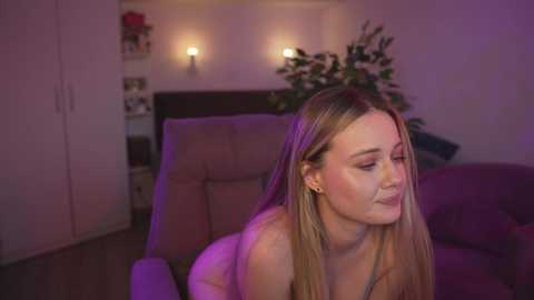 Media: A video of a fair-skinned, long-haired woman with a medium build, wearing a black top, sitting in a dimly lit living room. The background features a brown armchair, a plant, and soft lighting.