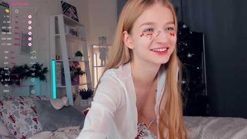 Media: Video of a smiling young woman with blonde hair, wearing a white shirt, lying on a bed adorned with festive pillows.