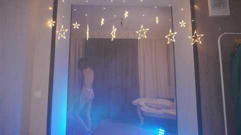 Media: Video of a dimly lit bedroom with a woman in a light dress facing away from the mirror, star-shaped fairy lights hanging above, and a bed with white sheets in the background.