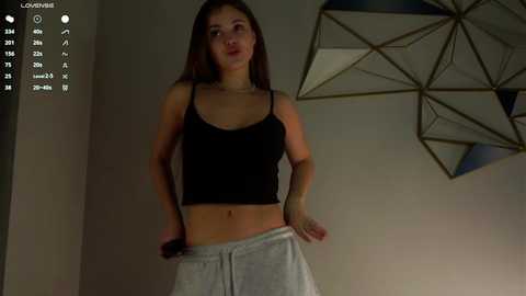 Media: A video of a slim, young Caucasian woman with long brown hair, wearing a black crop top and grey sweatpants, standing indoors.