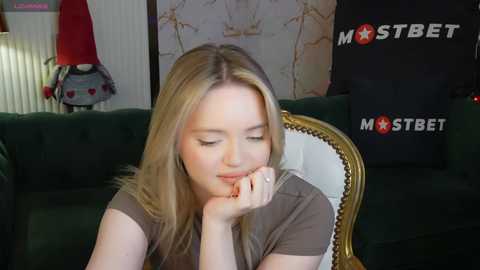 Media: Video of a blonde woman with fair skin, wearing a brown top, resting her chin on her hand, in a cozy living room with green couch and a decorative lamp.