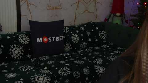 Media: Video of a dark green sofa with white snowflake patterns, adorned with a navy blue pillow featuring white text \"MST3K.\" Background shows a festive Christmas tree with red ornaments and a white wall with gold accents.