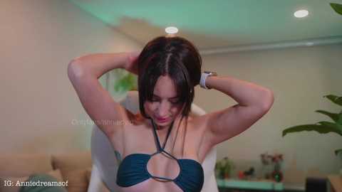 Media: Video of a slim, fair-skinned woman with dark hair, wearing a dark blue halter bikini, adjusting her hair in a modern living room with soft lighting.