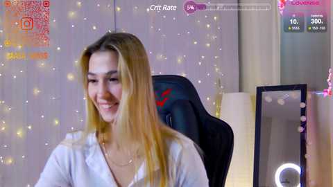 Media: Video of a smiling, light-skinned woman with long, blonde hair, wearing a white blouse, seated in a black gaming chair. Background features a soft, twinkling fairy light ambiance.