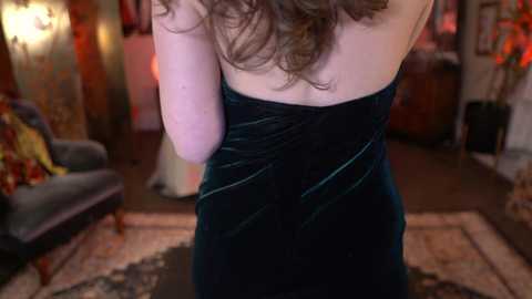 Media: Video of a woman with light skin, wavy brown hair, wearing a black velvet strapless dress, standing indoors with a cozy, dimly lit living room in the background.